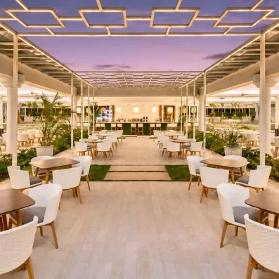 terrasse couverte restaurant tables grises carrées, chaises blanches, plantes partout et gazon au centre, bar au fond