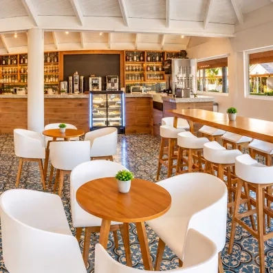 bar avec tables rondes en bois et chaises blanches, chaises hautes sur la droite, bar au fond pour petit déjeuner et gouter avec boissons chaudes, mais aussi bouteilles alcool