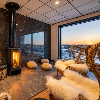 Espace détente du Mondi Club Sodankylä en Laponie avec poêle à bois, fauteuils en osier garnis de peaux de bête et baie vitrée vue sur le coucher de soleil.