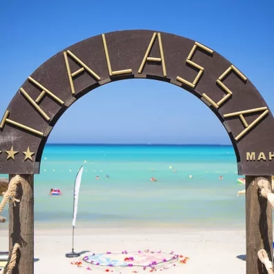 Arche en bois gravée au nom de l'hôtel Mondi Club Thalassa Mahdia avec vue sur la plage de sable blanc et la mer turquoise en Tunisie.