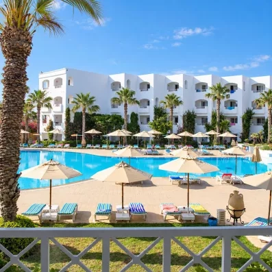 Grande piscine extérieure entourée de parasols, de transats et de palmiers devant le bâtiment principal de l'hôtel Thalassa Mahdia.