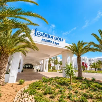 Façade blanche et entrée prestigieuse de l’hôtel Djerba Golf Resort & Spa sous un ciel bleu radieux.