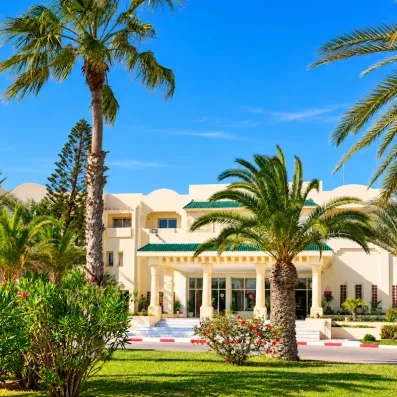 Façade blanche de l'hôtel Les Quatre Saisons Djerba entourée de palmiers sous un ciel bleu azur.