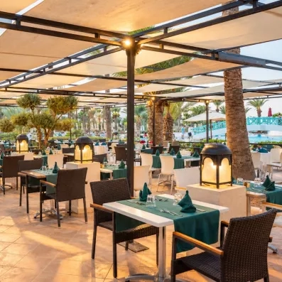 Un restaurant à ciel ouvert sous une pergola ombragée, entre palmiers et piscine, pour dîner dans une ambiance chaleureuse en fin de journée.