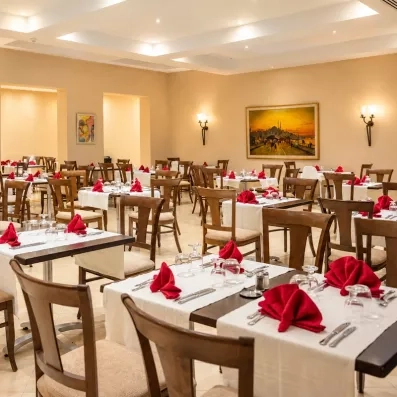 Une salle de restaurant élégante, aux tables dressées de nappes blanches et serviettes rouges, prête à accueillir les convives.