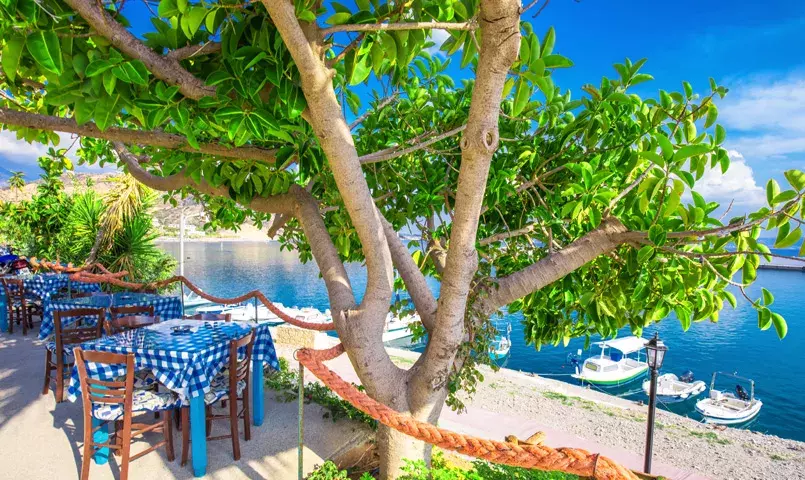 Terrasse de taverne en bord de mer en Crète, tables sous un arbre, bateaux au mouillage et eau turquoise, climat méditerranéen.