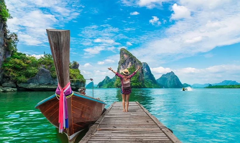 Une femme sur une jetée en bois, devant un bateau thaïlandais et de l'eau turquoise. 