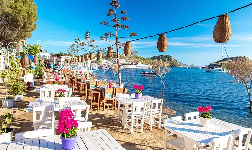 Terrasse de restaurant au bord de la mer avec tables blanches et vue sur des bateaux. 